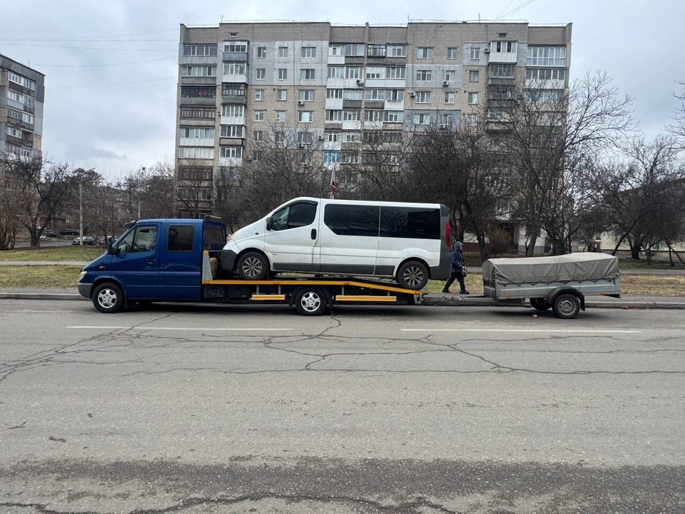 Синий эвакуатор перевозит белый микроавтобус, рядом прицеп