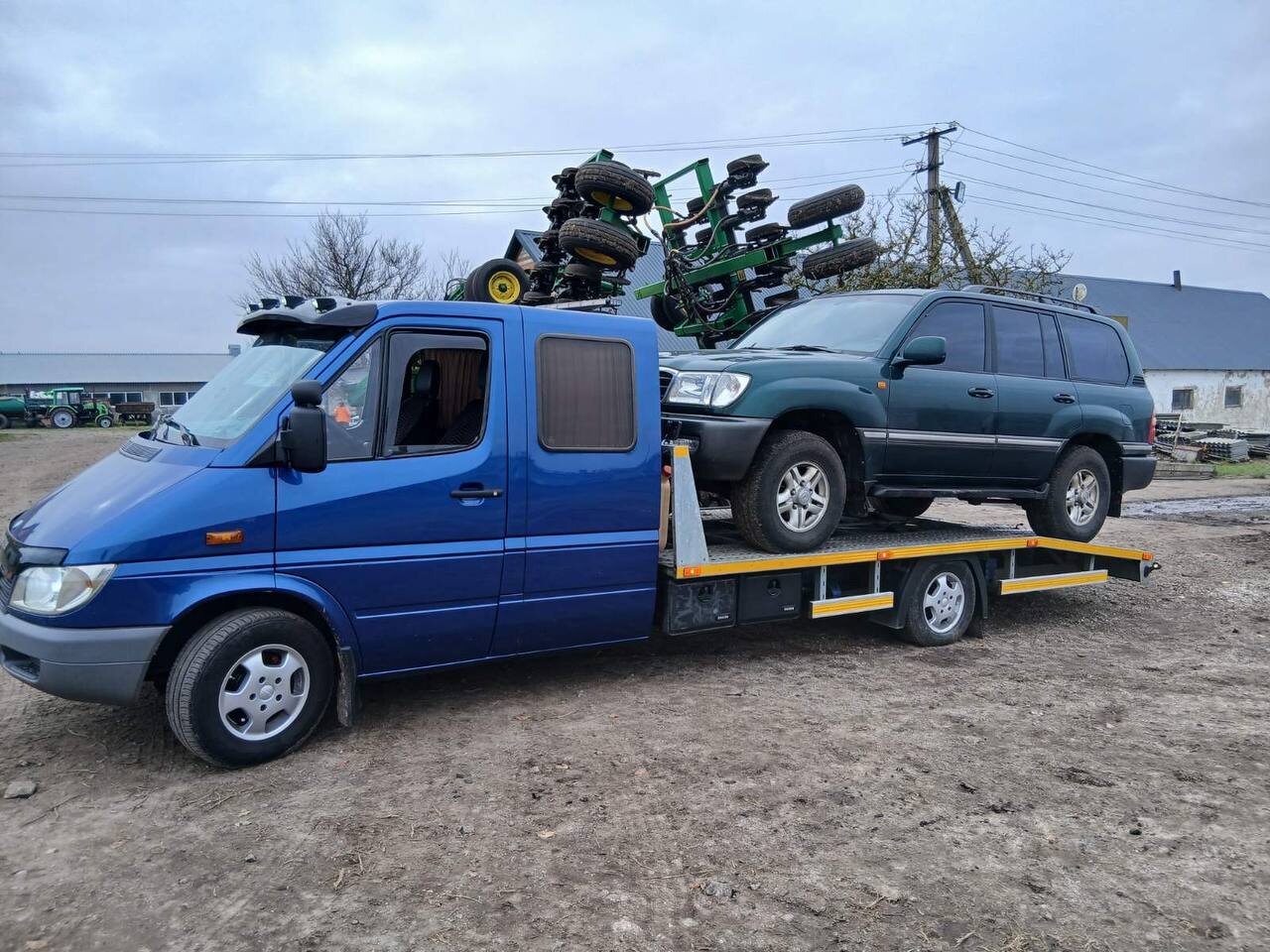 Синій евакуатор перевозить зелений позашляховик, поруч сільгоспобладнання