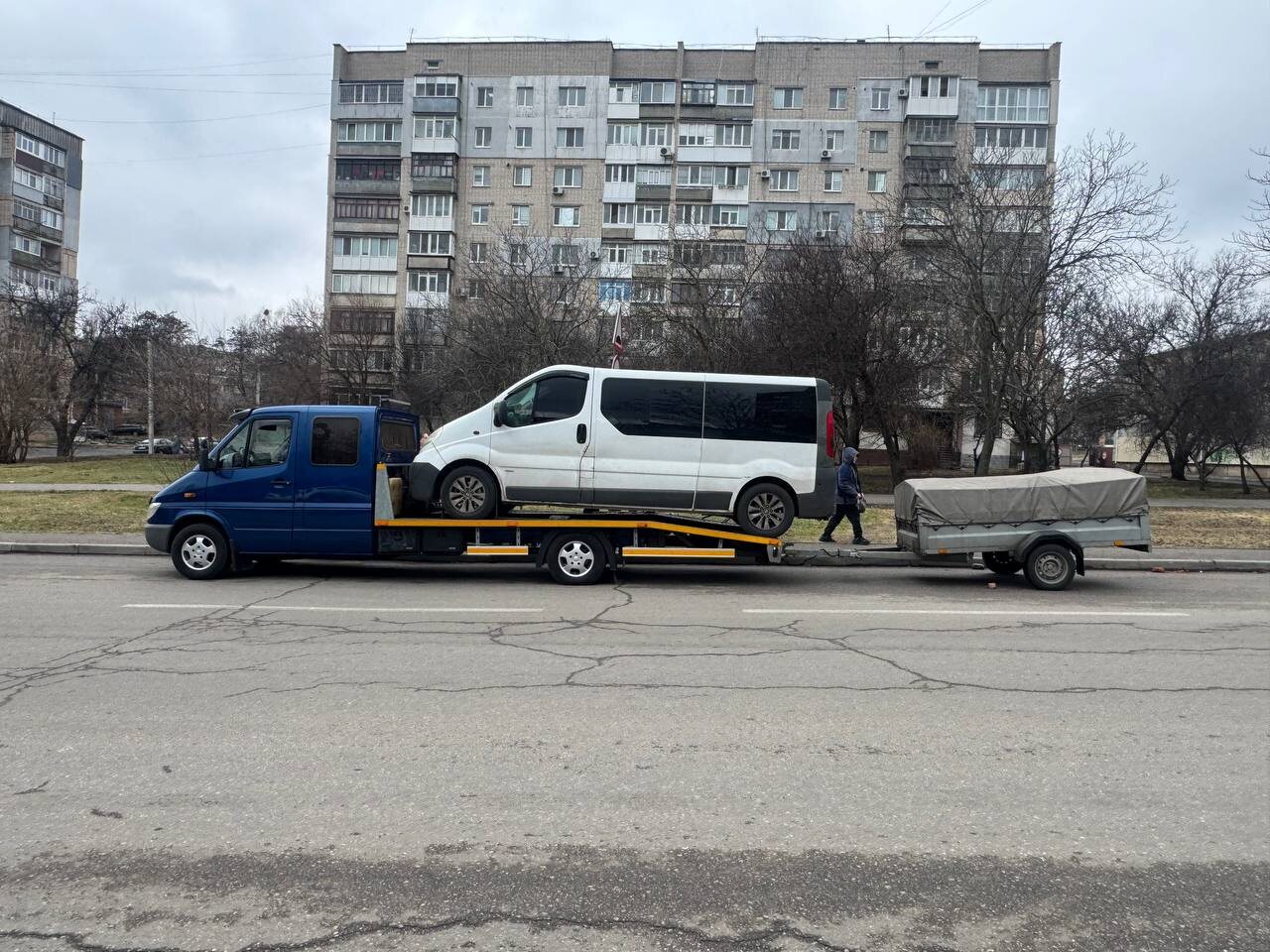 Синій евакуатор перевозить білий мікроавтобус, поруч причіп
